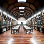 Fotografia de Candida Höfer intitulada IES Celanova I (2010), retratando uma biblioteca com estantes de madeira esculpidas, mesas centrais e iluminação simétrica.