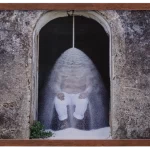Fotografia emoldurada retratando uma figura sentada sob uma cascata de pó branco, situada em uma entrada de arco com paredes de pedra envelhecidas. A obra contrasta a textura rústica do ambiente com a fluidez do material em movimento, criando uma atmosfera simbólica e introspectiva. A moldura de madeira adiciona um toque orgânico à composição.