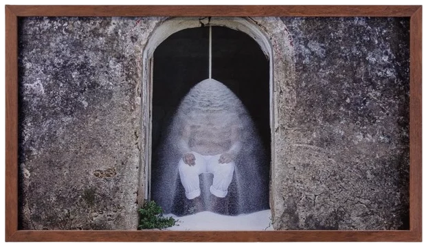 Fotografia emoldurada retratando uma figura sentada sob uma cascata de pó branco, situada em uma entrada de arco com paredes de pedra envelhecidas. A obra contrasta a textura rústica do ambiente com a fluidez do material em movimento, criando uma atmosfera simbólica e introspectiva. A moldura de madeira adiciona um toque orgânico à composição.