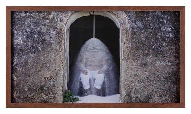 A framed photograph depicting a figure seated beneath a cascade of white powder, positioned within an arched entrance with weathered stone walls. The piece contrasts the rustic texture of the environment with the fluidity of the falling material, creating a symbolic and introspective atmosphere. The wooden frame adds an organic touch to the composition.