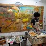 Artista Felipe Rezende em seu ateliê criando pinturas da exposição Arena, com cenas agrícolas sobre lona e materiais diversos.