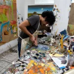 Artista Felipe Rezende em seu ateliê criando pinturas da exposição Arena, com cenas agrícolas sobre lona e materiais diversos.