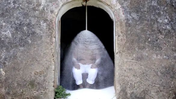 Scene from the video "Refino #2" (2017) by Tiago Sant’Ana, exhibited in the "Delírio Tropical" exhibition at the Pinacoteca do Ceará. The image shows a Black man sitting beneath a continuous cascade of sugar pouring over his body, set against the ruins of the former Engenho de Oiteiro in the Recôncavo of Bahia. The artwork reflects on coloniality and the enduring marks of slavery on racialized bodies.