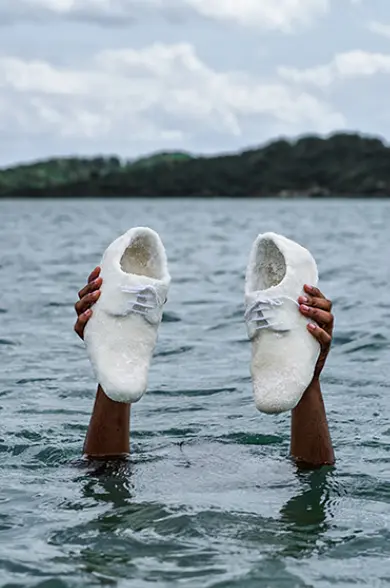 Hands emerge from the water holding a pair of white shoes made of sugar. Tiago Sant’Ana’s artwork symbolizes memory, displacement, and the lasting impact of slavery in the African diaspora. The background features a natural landscape with a cloudy sky and vegetation, creating a contrast between the fragility of the material and the strength of the historical message.